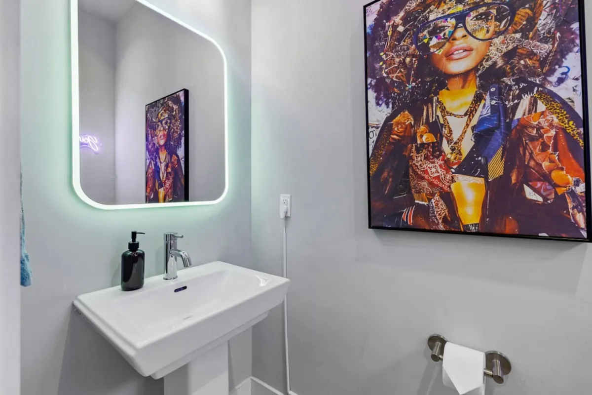 A chic bathroom featuring a large illuminated mirror, colorful modern artwork, and a contemporary sink setup. The energetic vibe is perfect for self-care routines or getting ready for a fun night out.
