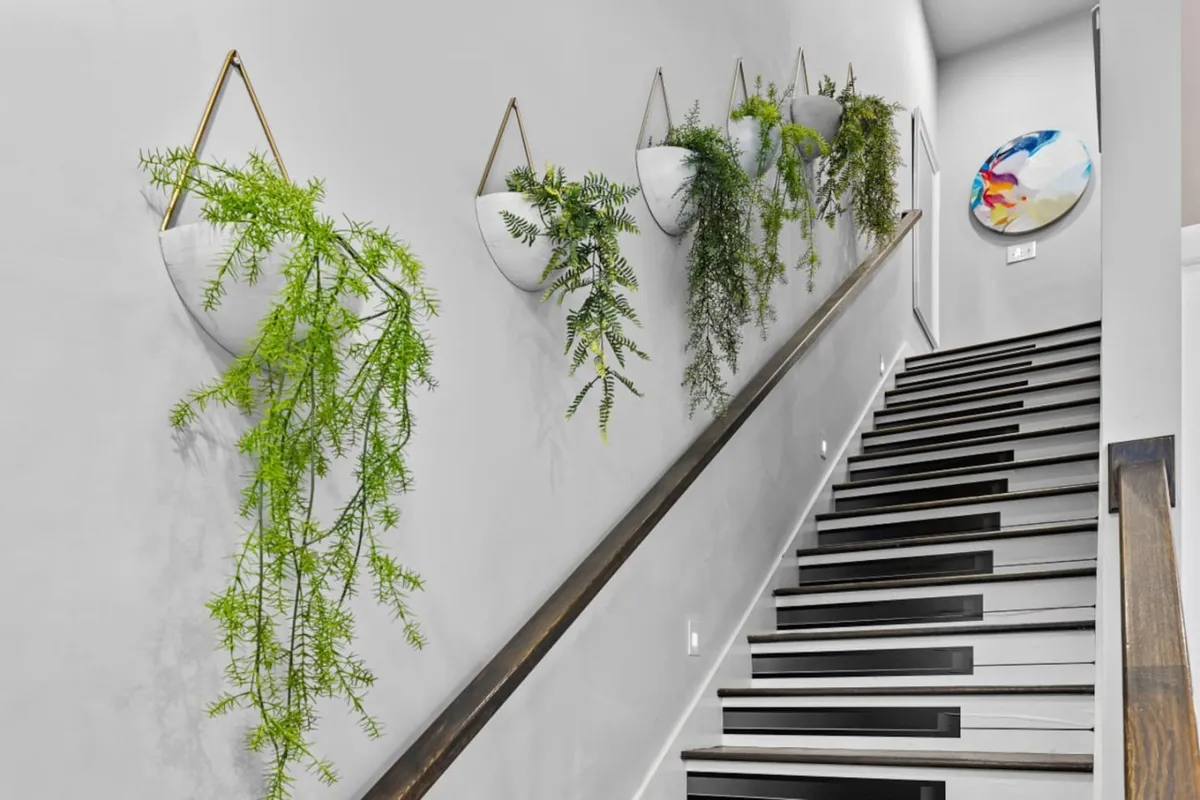A modern entryway or open area featuring hanging plants, a staircase, and eye-catching artwork. The scene suggests movement through the home, preparing to step out, or appreciating stylish touches before an evening out.