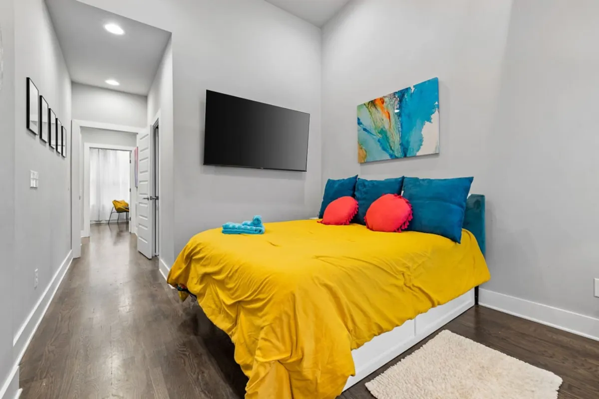 A brightly lit bedroom featuring a queen-sized bed dressed in a vivid yellow bedspread, colorful pillows, a modern abstract artwork above the headboard, and a flat-screen Smart TV on the opposite wall. The room exudes comfort and cheerfulness, ideal for restful nights or relaxing mornings.