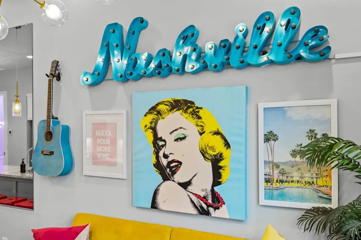 A vibrant living space featuring a prominent 'Nashville' neon sign, a blue guitar leaning by a yellow couch, and Marilyn Monroe artwork, all set against playful décor. The room exudes a sense of musical inspiration and serves as a social hub for guests to gather before or after nights out.