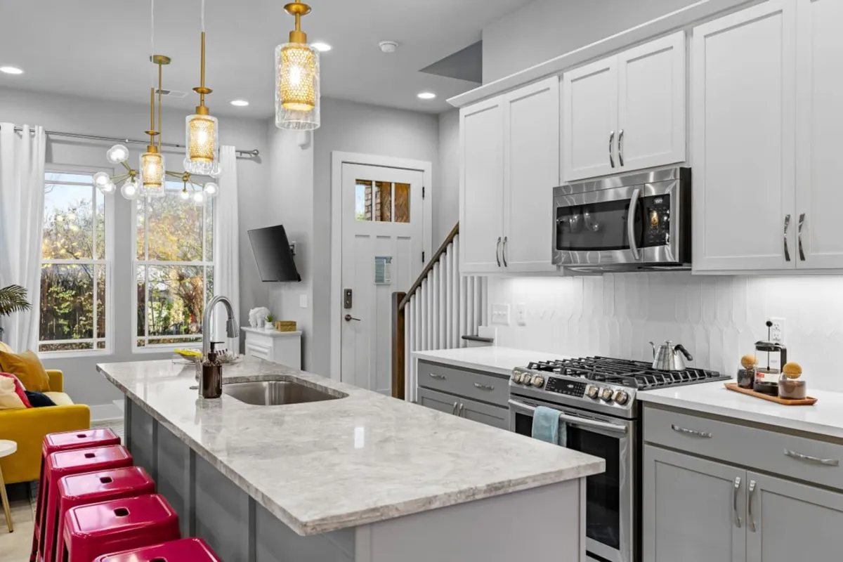 A modern kitchen with a spacious island, stainless steel appliances, colorful stools, and bright pendant lights. The open layout is designed for cooking, hosting, or gathering with friends and family, offering both functionality and style.
