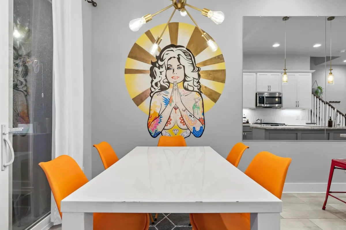 A vibrant and stylish dining area featuring bold orange chairs around a chic table, playful wall art—possibly a nod to Dolly Parton—and a modern, open layout perfect for gathering with friends. The setting is lively, inviting, and designed for memorable meals and celebrations.