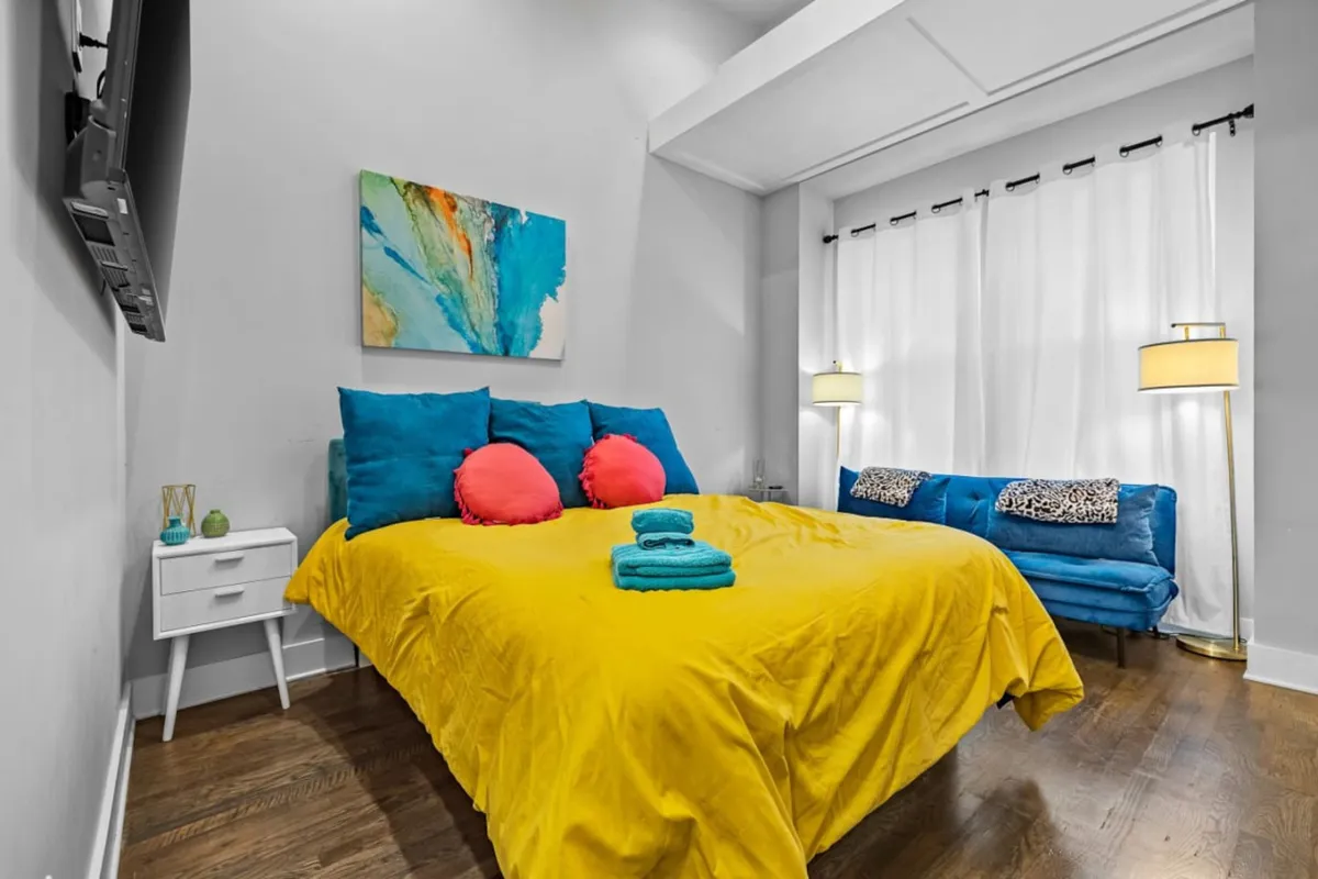 This lively living area features a bright yellow bedspread, colorful throw pillows, a comfy sofa with animal print, and eye-catching abstract wall art. The vibrant decor and casual seating invite relaxation and socializing, making it a perfect backdrop for group gatherings or unwinding after a night out.